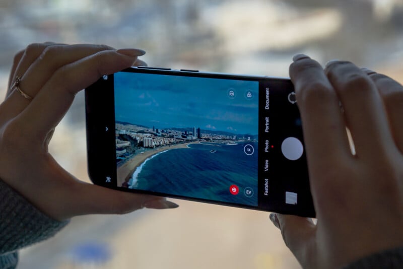 A person holds a smartphone, capturing a scenic photo of a coastal city through a window. The phone screen shows a beach, ocean, and city skyline under a cloudy sky. Hands with polished nails frame the device.