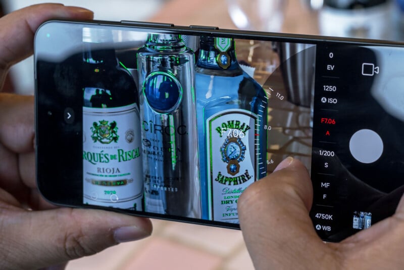 A person is using a smartphone camera to take a photo of three bottles: Marques de Riscal Rioja wine, Ciroc vodka, and Bombay Sapphire gin. The camera interface, with various settings and controls, is visible on the screen.