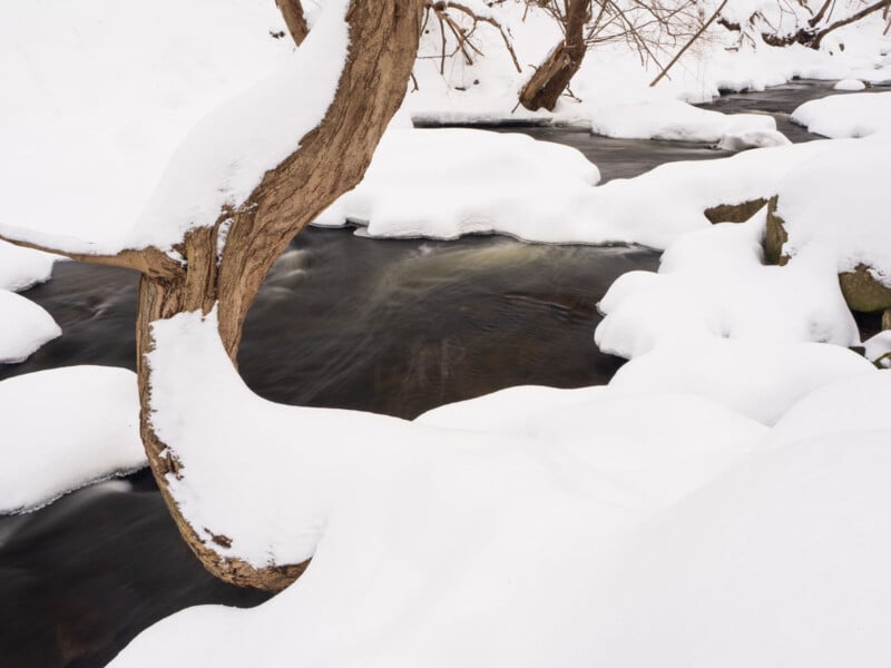 A snow-covered stream winds through a winter landscape. A tree trunk arches over the water, surrounded by rocks and snowy banks. The scene is serene and calm, with soft light highlighting the fresh snow.