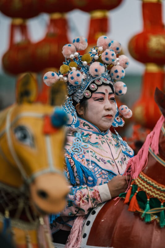 Person wearing ornate traditional costume and headpiece featuring colorful pompoms and intricate patterns, riding a decorative horse figure. Red lanterns hang in the background, creating a festive atmosphere.