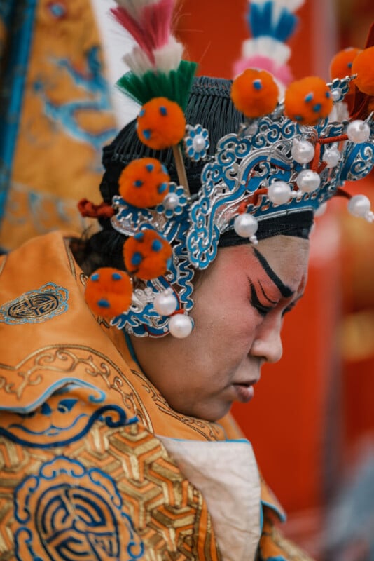 A person in ornate traditional attire, adorned with an elaborate headdress featuring colorful pom-poms and pearls, is seen in profile. The detailed clothing and makeup suggest a cultural or theatrical event.