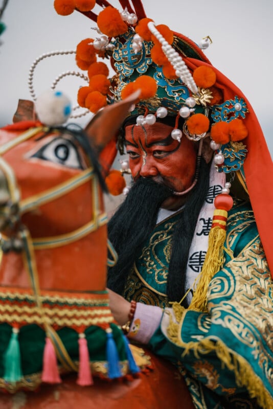 A person dressed in traditional Chinese opera costume with ornate headgear and face paint is seated on a decorated horse figure. The outfit features intricate embroidery and vivid colors, including red, green, and gold.