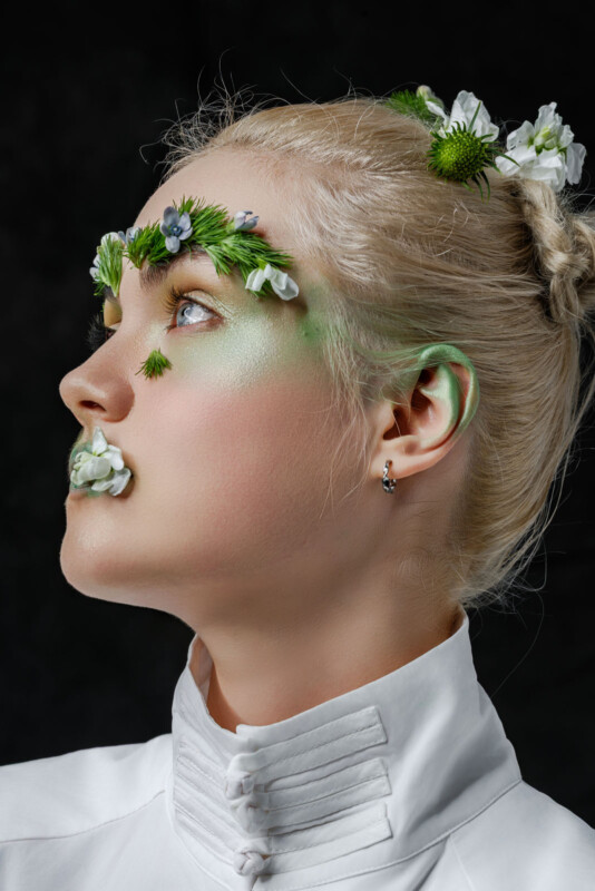 A person with platinum blonde hair styled elegantly wears a white high-collared top. Green and white floral elements adorn their eyebrows, lips, and hair, creating an artistic, nature-inspired look against a dark background.