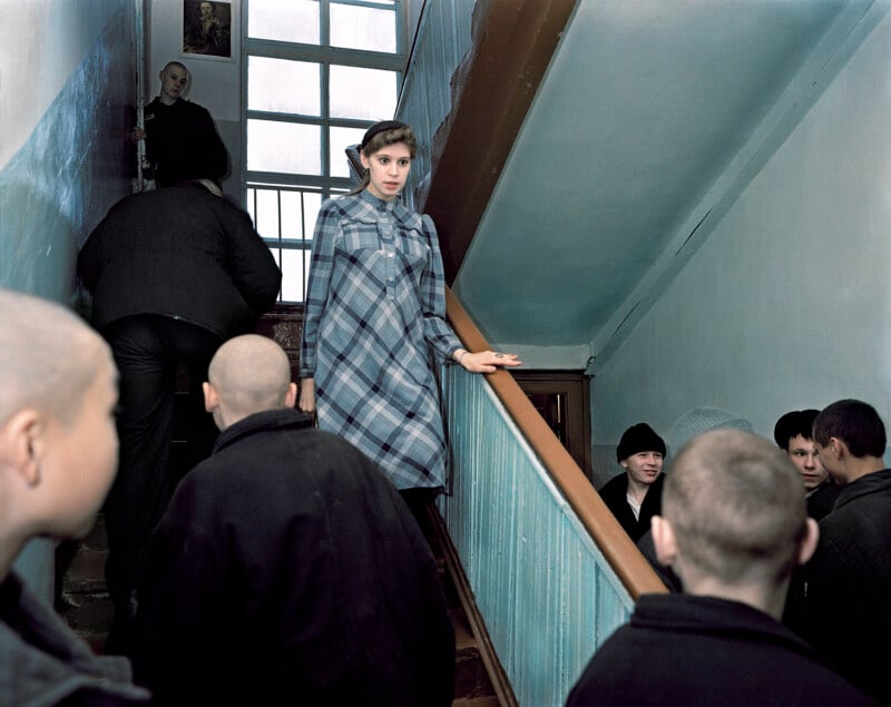 A young woman in a plaid coat descends a staircase in an old building. She is surrounded by several people with shaved heads wearing dark clothing. The stairwell has a large window and worn blue-green walls.