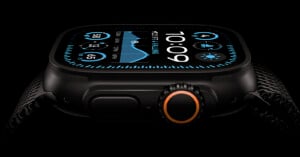 Close-up image of a smartwatch with a dark body and an intricate strap. The display shows time, weather, and activity metrics. A prominent orange-accented dial is visible on the side against a black background.