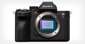 A Sony Alpha 7R camera with a visible E-mount sensor. The camera body is black with textured grip on the left side and control dials on top. The brand logo is shown on the viewfinder area in white.