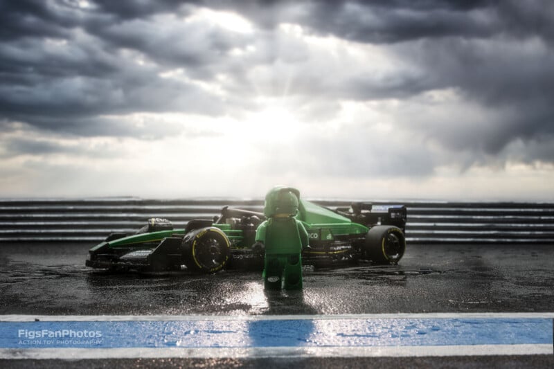 A toy figure in a green racing suit stands on a wet race track facing a green formula-style race car. Dark clouds loom above, with dramatic sunlight breaking through, reflecting on the wet track. The scene captures a cinematic and intense atmosphere.