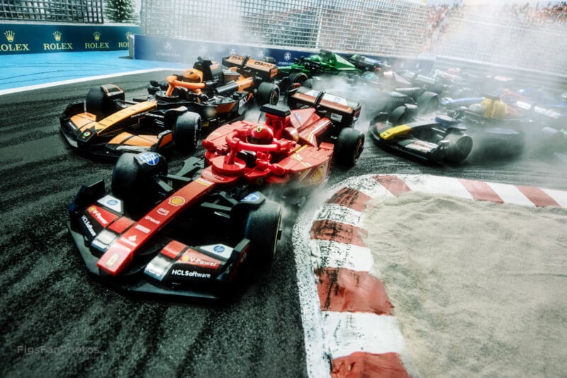 Formula 1 cars aggressively navigate a turn on a racetrack, with a red car leading the pack. There is visible tire smoke, and the grandstand in the background shows a crowd of spectators watching closely.