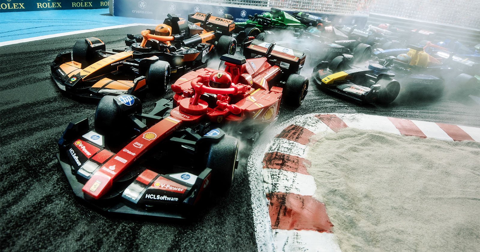 A group of small, toy-like racing cars on a track, taking a sharp corner. The lead car is red, followed closely by orange and black cars. Smoke trails from the tires, and a sandy section is visible on the right.