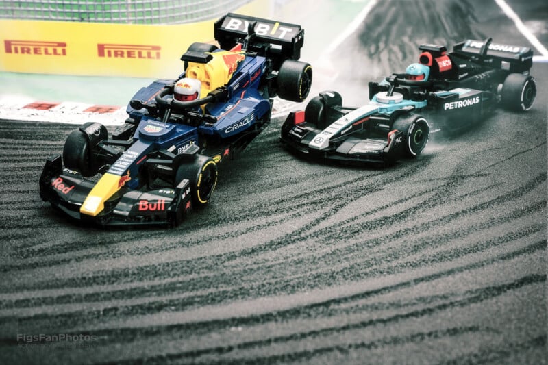 Two toy Formula 1 cars race on a track. One car is black with white and blue accents, and the other is black with blue and red accents. The track has visible skid marks, and Pirelli advertisements are in the background.