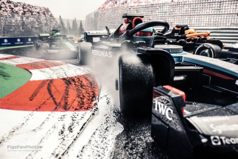 A close-up shot of toy Formula 1 race cars navigating a wet, curving track. Water sprays dramatically from the tires as they speed around the corner. The scene captures the intensity and excitement of a high-speed race.