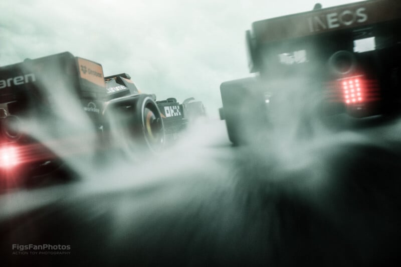 Low-angle shot of two Formula 1 cars racing on a wet track, with spray from the tires creating a misty effect. The overcast sky adds to the dramatic atmosphere.