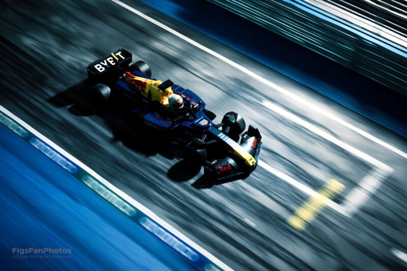 A Formula 1 car speeds down a well-lit racetrack at night. The driver is wearing a helmet, and the car displays multiple sponsor logos. Motion blur emphasizes the high speed.