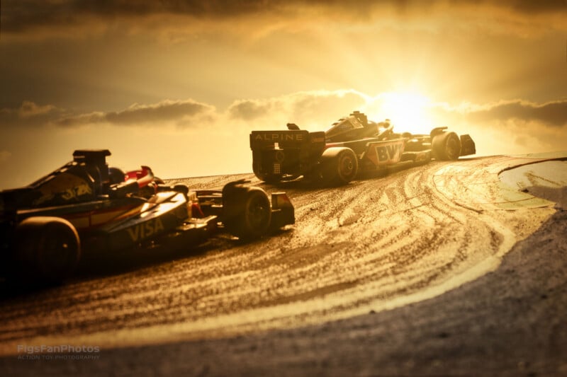 Two Formula 1 cars race on a track with a dramatic sunset in the background. The sky is filled with warm, golden light, casting long shadows across the track. One car is ahead, both showing sponsor logos. Tire marks are visible on the track.