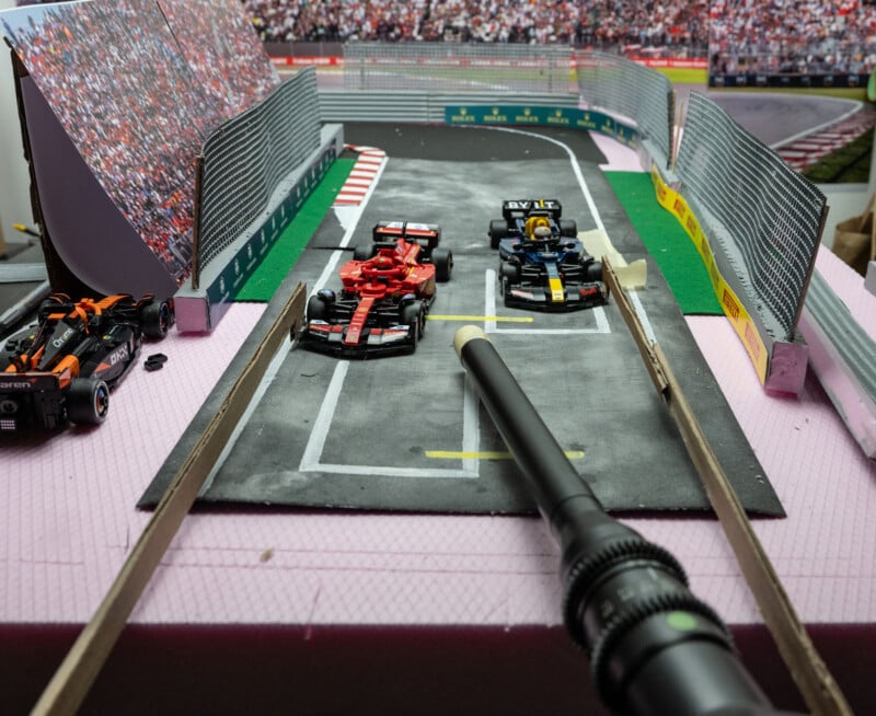 A diorama of a Formula 1 pit lane shows model cars on tracks. Two cars are in the pit box area, with crowd stands and safety barriers around. A close-up camera lens is positioned in the foreground, capturing the scene.