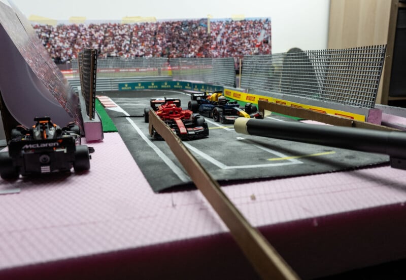 Model Formula 1 cars on a miniature racetrack, with a crowd backdrop. The scene includes a McLaren car and other F1 models lined up. Various tools and materials are visible, suggesting setup or construction.