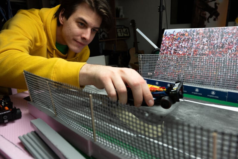 A person in a yellow hoodie is carefully arranging a model race car on a small-scale racetrack. The setup includes fencing and a printed backdrop of a cheering crowd. A guitar is visible in the background.