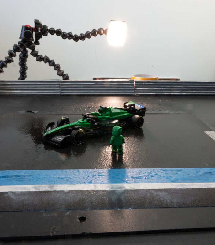 A miniature green race car and a green toy figure are on a black surface with blue and white stripes. A bright light on a flexible stand illuminates the scene, creating reflections on the surface.