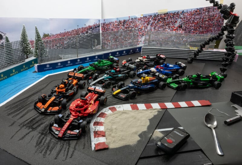 A detailed miniature Formula 1 race diorama featuring several race cars on a track. The scene includes a sand trap, crowd background, and fencing. A camera lens and Gorillapod are visible on the edge, capturing the setup.