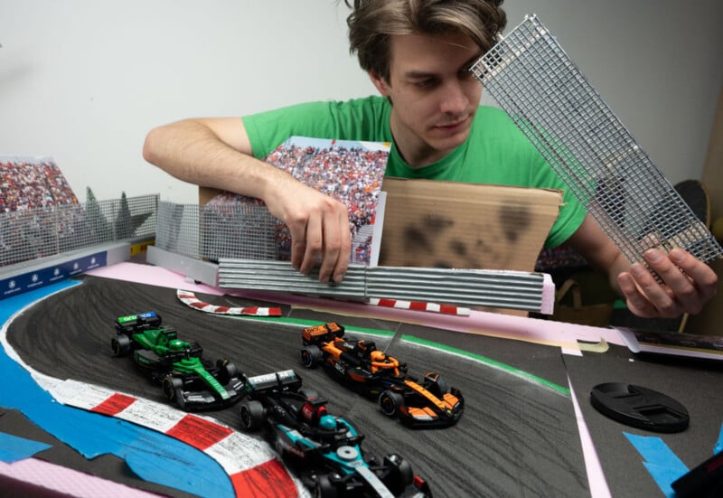 A man in a green shirt builds a miniature race car track with model cars on it. He is holding a small section of a wire fence. The track is surrounded by stands filled with tiny spectators, depicting a lively racing scene.