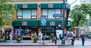 Street view of a busy storefront with "B&H" signage on awnings. People are walking on the sidewalk, and trees and potted plants decorate the scene. Traffic signs point to East 495/West 34th St. It's a bustling urban environment.