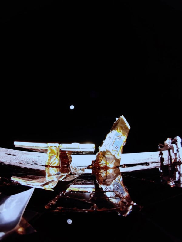 A dark space scene with a shiny spacecraft component reflecting light. In the background, Earth appears as a small, distant sphere against the blackness of space. The foreground includes metallic and golden materials on the spacecraft.