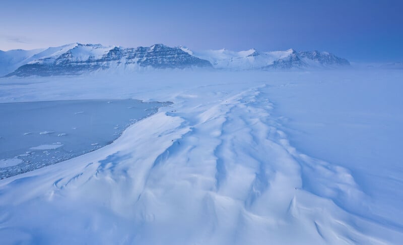 A vast icy landscape with snow-covered ridges in the foreground and a frozen body of water to the left. Rugged mountains rise in the background under a clear, pale blue sky. The scene conveys a sense of remote, winter tranquility.