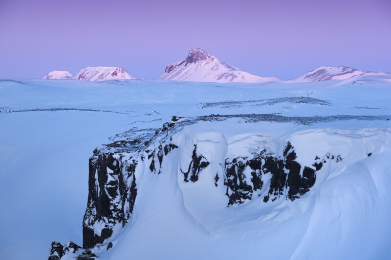 A snow-covered landscape with a rocky cliff in the foreground and several distant mountains under a purple sky at dawn or dusk. The scene is serene, depicting a winter environment with soft lighting and expansive snowfields.