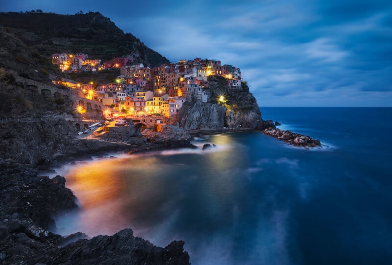 A coastal village with brightly lit buildings perched on a rocky cliff at dusk. The ocean waves gently crash against the rocks, and the sky is a moody blue with scattered clouds. The scene exudes a tranquil, picturesque atmosphere.