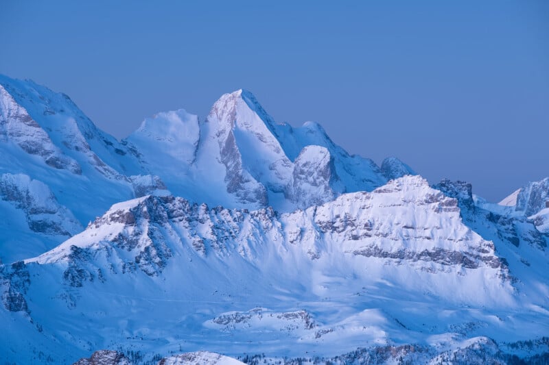 Snow-covered mountain peaks under a clear blue sky, with soft evening light enhancing the landscape's serene and majestic appearance.