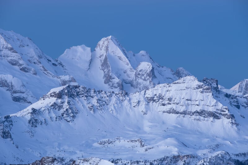 Snow-covered mountains under a clear blue sky, with jagged peaks rising prominently. The slopes are blanketed in thick, white snow, creating a serene and majestic winter landscape.