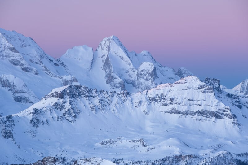 Snow-covered mountain range under a purple and blue gradient sky at dusk, showcasing jagged peaks and ridges with soft evening light.