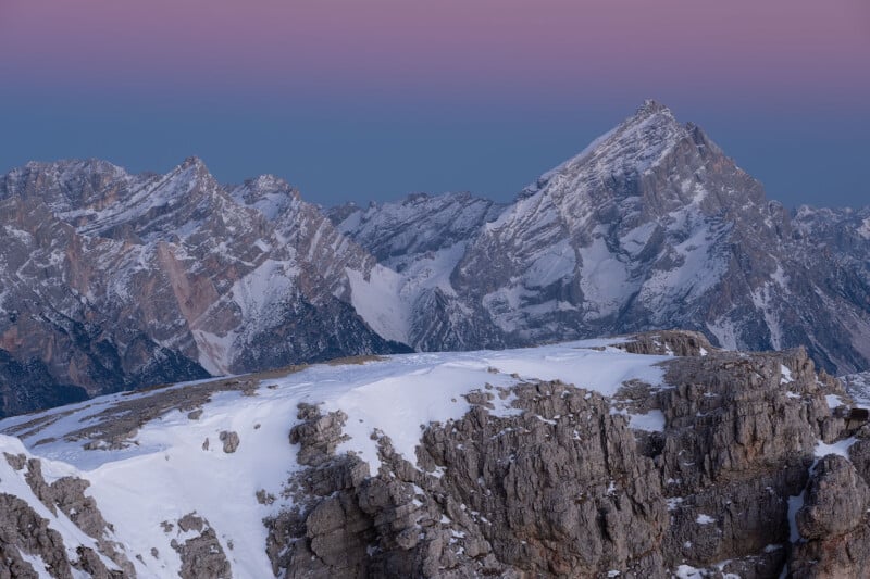 Snow-covered mountain peaks under a twilight sky transition from pink to blue hues. Rocky foreground and distant ridges add depth to the serene, crisp alpine scene.