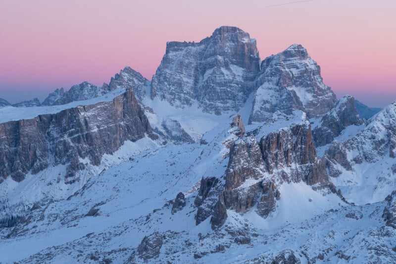 Snow-covered mountains under a pink and blue sky during sunrise. The rugged peaks and ridges are highlighted by the soft morning light, creating a serene and majestic landscape.