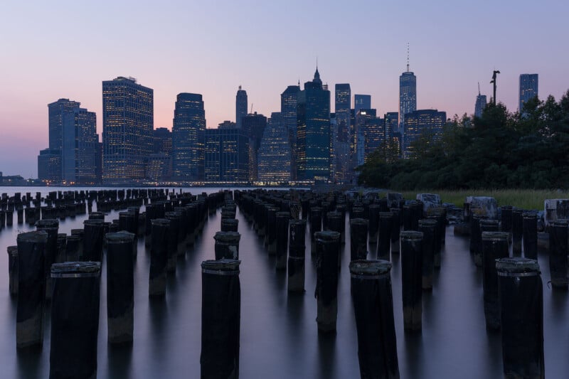 A serene evening view of Manhattan's skyline from Brooklyn, with silhouetted skyscrapers and a pink sky. Wooden pilings in the foreground stretch into the East River, adding depth to the tranquil scene.