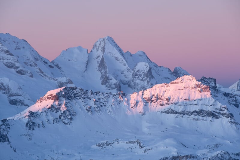 Snow-covered mountain peaks bathed in warm pink and purple hues during sunset or sunrise, creating a serene and majestic winter landscape.