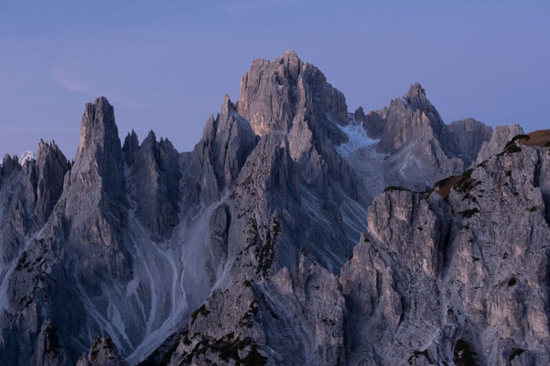 Majestic jagged mountain peaks under a clear blue sky in the early evening. The rocky formations have sharp edges and varied textures, with subtle shadows enhancing their rugged appearance. Snow patches cling to some areas.