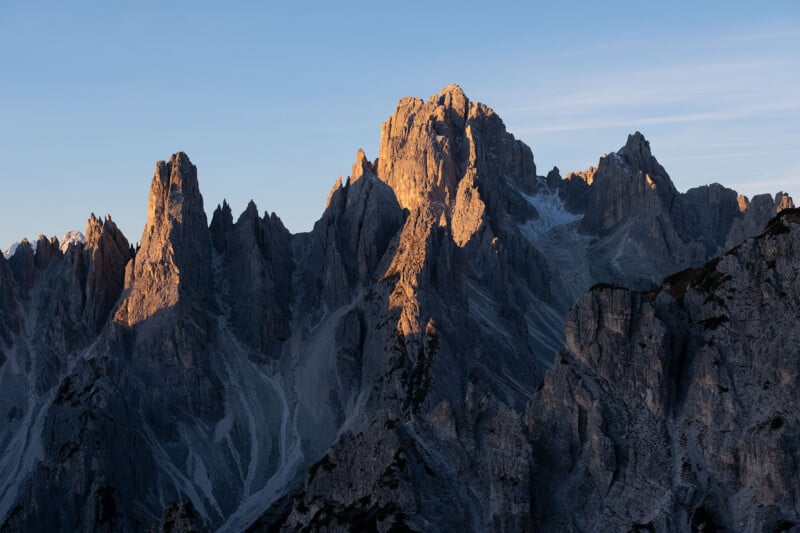 Sunlight bathes the jagged peaks of a mountain range, highlighting rugged textures and shadows. The clear sky in the background adds a serene contrast to the imposing, rocky formations.