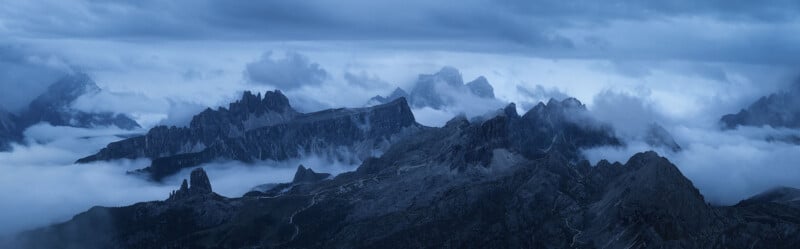 A panoramic view of a mountain range shrouded in mist and clouds at twilight. The rugged peaks and ridges are silhouetted against the dusky sky, creating a dramatic and serene landscape.