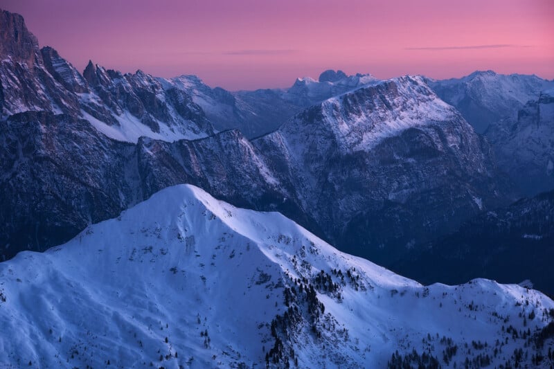 Snow-covered mountains under a purple and pink sky at sunset. The jagged peaks and ridges form a dramatic landscape, with soft light highlighting the snow. Forested areas are visible near the base of the mountain range.