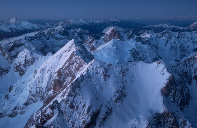 Aerial view of snow-capped mountain peaks under a clear twilight sky, with rugged terrain and a subdued blue hue enveloping the landscape. The distant horizon features additional mountain ranges fading into the background.