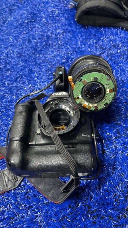 A disassembled camera lies on a blue carpet. The camera body is without a lens, which is placed nearby showing its internal circuit board. A camera strap is attached to the body.