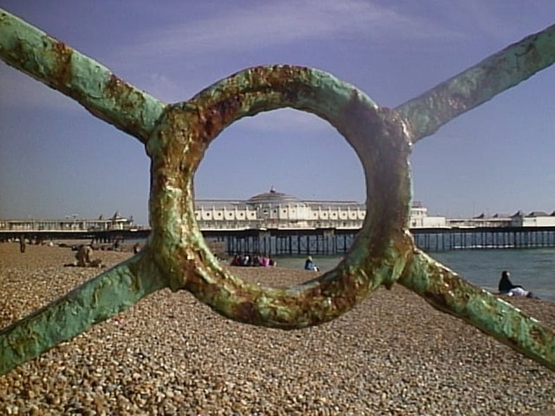 Rusty railing with a circular opening frames a view of a long pier extending over a pebble beach. The pier has a building with a domed roof. The sky is clear, and people are visible on the beach.