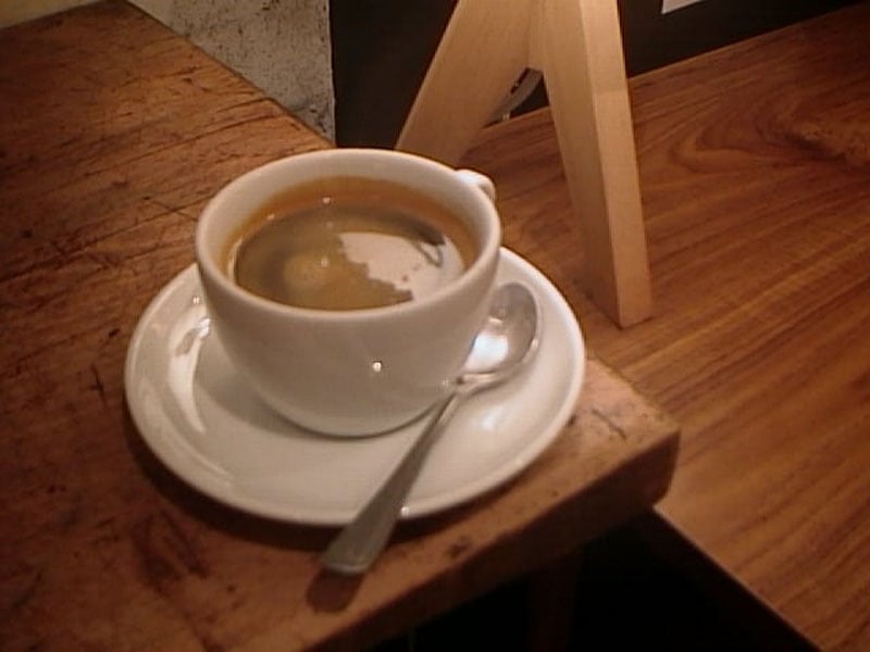 A cup of espresso sits on a saucer with a spoon, placed on a wooden table. The background includes part of a wooden easel or stand. The espresso has a smooth, dark surface with some crema visible.