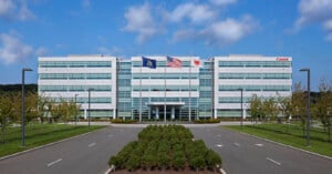 A modern, four-story office building with reflective glass windows. Two flagpoles display an American flag and another flag. Trees and a landscaped median line the road leading to the entrance under a partly cloudy sky.