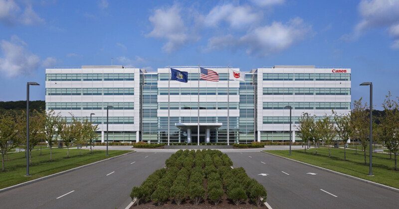 A modern, four-story office building with reflective glass windows. Two flagpoles display an American flag and another flag. Trees and a landscaped median line the road leading to the entrance under a partly cloudy sky.