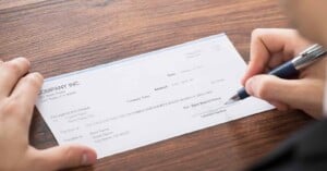 A person signs a check on a wooden table. The check, issued by "Company Inc.," is addressed to an individual, with a date of January 15, 2023. The amount is two hundred forty-seven dollars. A pen is held in the person's right hand.