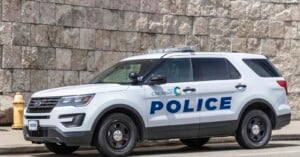 A Cincinnati Police SUV is parked on the street next to a stone wall and fire hydrant. The vehicle is white with blue lettering that says "POLICE" and a logo indicating it's from Cincinnati.