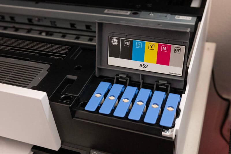 Close-up of an open printer showing five ink cartridges in a row. The cartridges are color-coded and labeled: black (BK), photo black (PB), cyan (C), magenta (M), and yellow (Y). The printer model is labeled as 552.