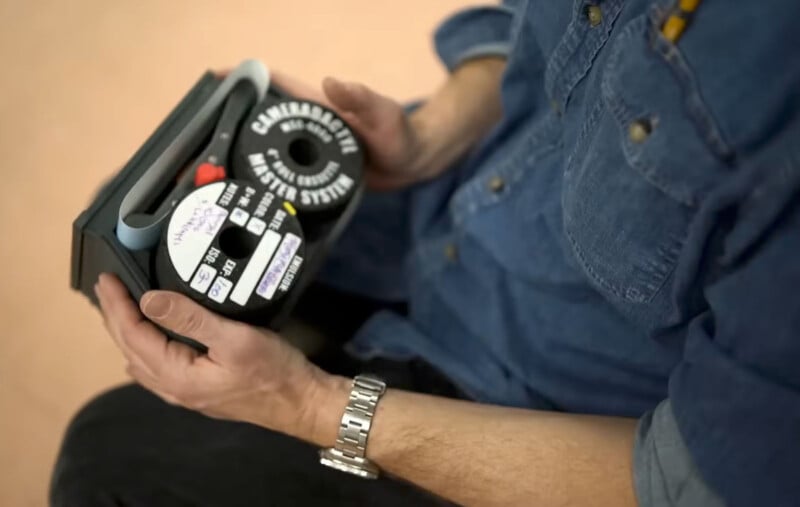 A person in a denim shirt holds an unusual cassette labeled "Cambracase System." The cassette has multiple labels and a red protrusion. The individual is seated, wearing a silver wristwatch.